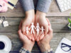Symbolbild für das Portfolio einer Kranken- und Unfallversicherung: Hände halten Familie in Form von Papierfiguren. Marketing-Analysen