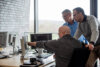 Drei Software Developer stehen vor Monitoren und diskutieren Software Herausforderungen, Licht von hinten, grosses Fenster, Backsteinwand